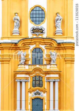 The ornate yellow and white baroque facade of Melk Abbey, adorned with sculptures and decorative windows, reflecting the grandeur of the historic monastery. Austria The ornate yellow and white baroque facade of Melk Abbey, adorned with sculptures and decorative windows, reflecting the grandeur of the historic monastery. Austria 113028016
