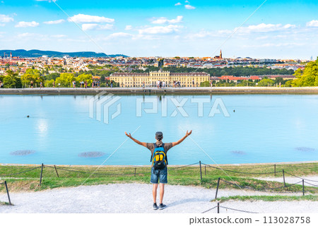 A traveler with raised arms stands before the vast Schonbrunn Palace and its Great Parterre, overlooking the ornamental gardens, tranquil water, and city skyline on a sunny day. Vienna, Austria A traveler with raised arms stands before the vast Schonbrunn Palace and its Great Parterre, overlooking the ornamental gardens, tranquil water, and city skyline on a sunny day. Vienna, Austria 113028758