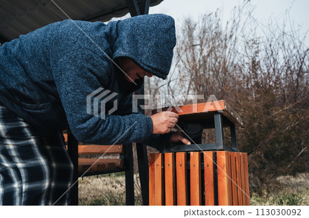 homeless elderly old Caucasian man rummages for food and garbage in trash can in a park in autumn 113030092