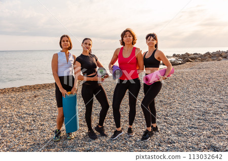 Group of four Caucasian pretty adult women holds sports mats on pebble beach. Sea and wild coast in background. Fitness outdoor training Group of four Caucasian pretty adult women holds sports mats on pebble beach. Sea and wild coast in background. Fitness outdoor training 113032642
