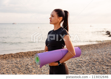 Outdoor activity. Portrait of young Caucasian woman holds sport mat and posing on beach. Copy space. Concept of wellness Outdoor activity. Portrait of young Caucasian woman holds sport mat and posing on beach. Copy space. Concept of wellness 113032646