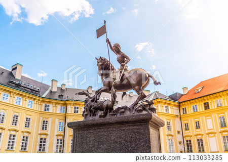 The bronze statue of Saint George triumphantly slays a dragon, set against the vibrant backdrop of Prague Castle under a blue sky. Prague, Czechia 113033285