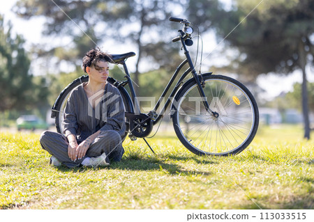 A woman sits on the grass next to a black bicycle A woman sits on the grass next to a black bicycle 113033515