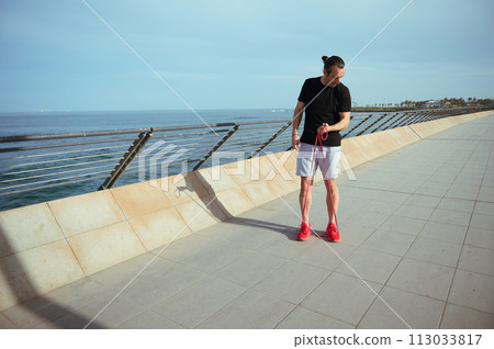 Full length shot of a man with athletic body doing exercises on arms, pumping up biceps using a resistance band, standing on the promenade against Atlantic ocean background. Active people and sport 113033817