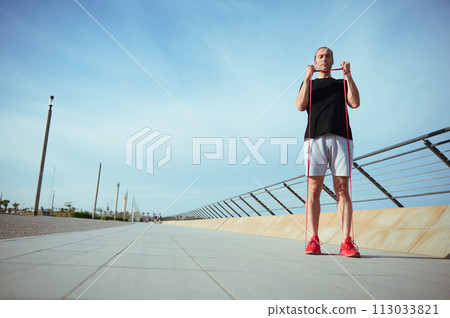 Man with athletic body performs bodyweight fitness exercises using a resistance band. Muscular guy in black t-shirt, gray sports shorts and red sneakers working out outdoors. Strength and motivation Man with athletic body performs bodyweight fitness exercises using a resistance band. Muscular guy in black t-shirt, gray sports shorts and red sneakers working out outdoors. Strength and motivation 113033821