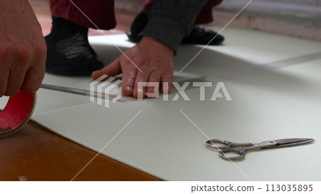 male hands glue together with transparent tape sheets of soft light-colored underlay for laminate on the floor and scissors lying nearby, do it yourself preparing the flooring at home 113035895