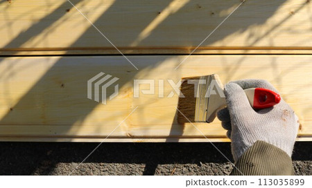 a hand in a fabric glove holds a paint brush and moves a layer of light brown paint over the surface of a board of fresh wood to impregnate and protect the wooden building material 113035899