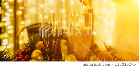 Two champagne glasses filled with champagne are placed on a table with a fruit arrangement. The glasses are surrounded by lights, creating a festive atmosphere. 113036270