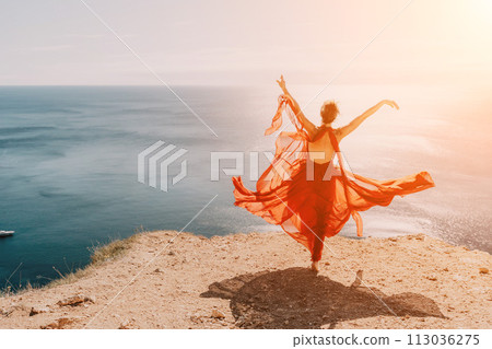 Woman in red dress on sea. Side view a Young beautiful sensual woman in a red long dress posing on a rock high above the sea on sunset. Girl on the nature on blue sky background. Fashion photo. Woman in red dress on sea. Side view a Young beautiful sensual woman in a red long dress posing on a rock high above the sea on sunset. Girl on the nature on blue sky background. Fashion photo. 113036275