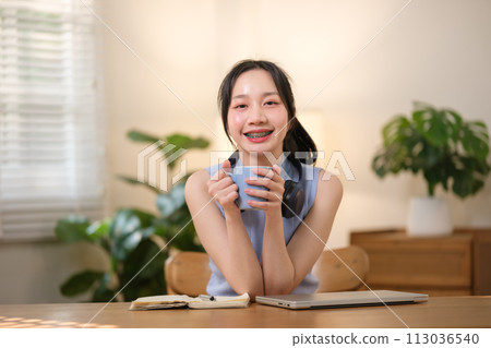 Young attractive happy Asian female student holding a cup of coffee sitting in the living room smiling and looking at the camera while working on a laptop at the home office. 113036540