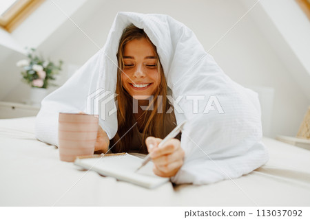 A woman, happy and smiling, lies on a bed under a blanket, writing in a notebook A woman, happy and smiling, lies on a bed under a blanket, writing in a notebook 113037092