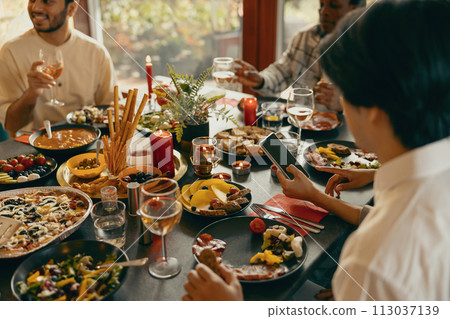 Close up of friends sitting at festive dinner table at home, drinking wine and looking at phone 113037139