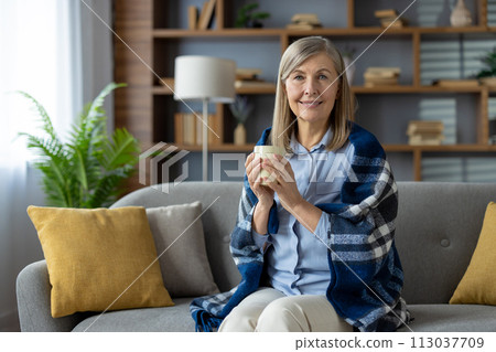 A relaxing scene of a woman sitting on a couch, covered with a blanket, and holding a coffee mug in a well-decorated living room. A relaxing scene of a woman sitting on a couch, covered with a blanket, and holding a coffee mug in a well-decorated living room. 113037709