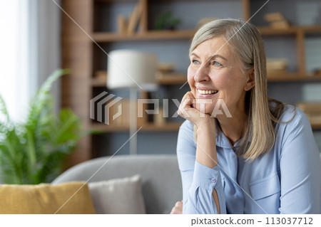 A contemplative adult woman sits on a couch, surrounded by soft, cozy pillows, lost in thought in a serene living room setting. 113037712