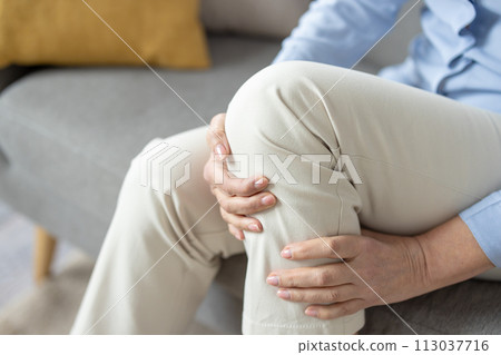 Close-up image of a mature woman sitting on a couch and holding her knee, depicting pain or discomfort in the joint. Close-up image of a mature woman sitting on a couch and holding her knee, depicting pain or discomfort in the joint. 113037716