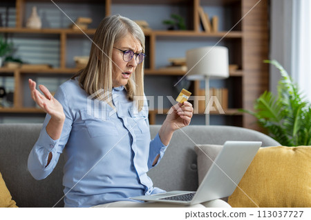 Mature woman sitting on the couch looking puzzled with a credit card and a laptop, possibly dealing with online shopping or banking issues. Mature woman sitting on the couch looking puzzled with a credit card and a laptop, possibly dealing with online shopping or banking issues. 113037727