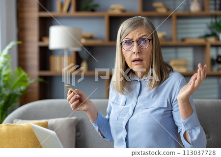 Adult woman sitting at home, holding a credit card with a puzzled expression as she looks at a laptop screen. Adult woman sitting at home, holding a credit card with a puzzled expression as she looks at a laptop screen. 113037728