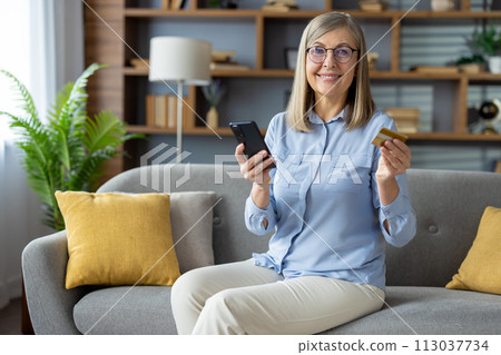 Modern consumerism concept with a woman holding credit card and smartphone for online shopping while sitting comfortably on a sofa at home. 113037734