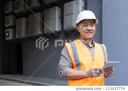 Portrait of a smiling young male construction worker in a hard hat and vest holding a tablet in his hands and looking at the camera. 113037774