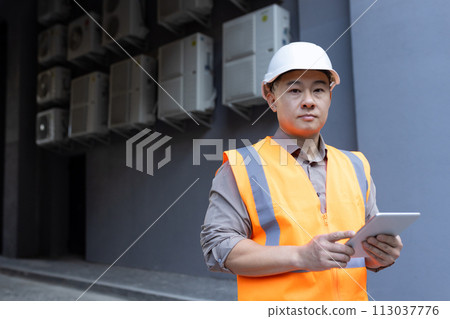Close-up portrait of a young Asian man wearing a hard hat and vest, standing outside a factory, construction site, holding a tablet and looking seriously into the camera. Close-up portrait of a young Asian man wearing a hard hat and vest, standing outside a factory, construction site, holding a tablet and looking seriously into the camera. 113037776