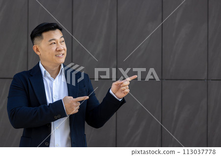Asian young male businessman standing near an office building on a gray wall background in a suit and smiling pointing with his fingers on a gray wall, blank background mockup. Asian young male businessman standing near an office building on a gray wall background in a suit and smiling pointing with his fingers on a gray wall, blank background mockup. 113037778