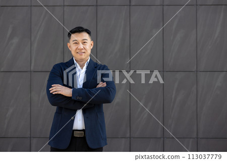 Portrait of young successful Asian businessman, lawyer in suit standing outside against wall background, arms crossed on chest and smiling confidently at camera. 113037779