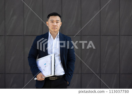 Portrait of a young successful serious young Asian man standing outside a business center against a wall, holding documents and an agreement in his hand. He looks seriously into the camera. 113037780