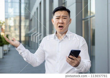 Close-up portrait of an angry young Asian man in a white shirt standing outside an office building, holding a phone and throwing his hands up in frustration and looking at the camera. 113037781