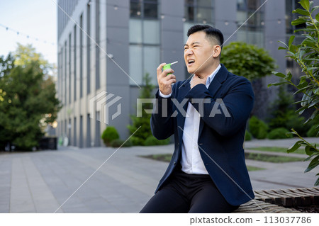 Suffering from a sore throat, a young Asian male businessman sits on a bench outside an office center and sprays medicine aerosol down his throat, holding his hand to his neck. Suffering from a sore throat, a young Asian male businessman sits on a bench outside an office center and sprays medicine aerosol down his throat, holding his hand to his neck. 113037786