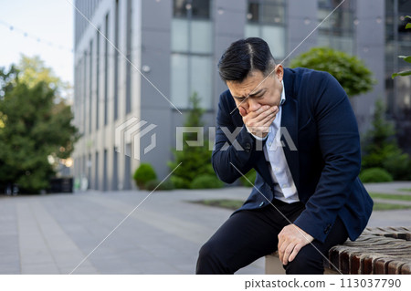Asian young man in a suit sits on a bench in the street with his hand covering his mouth, feels severe nausea and urge to vomit. Asian young man in a suit sits on a bench in the street with his hand covering his mouth, feels severe nausea and urge to vomit. 113037790