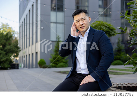 A young Asian man in a business suit is sitting on a bench on the street near an office center, holding his hand over his ear, grimacing in pain. 113037792