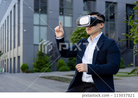 Serious and focused young man businessman sitting on a bench outside an office building wearing a virtual mask and working online. Serious and focused young man businessman sitting on a bench outside an office building wearing a virtual mask and working online. 113037821