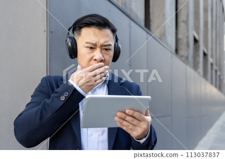A young Asian man in a suit and headphones is standing outside and is looking terrified at the screen of the tablet he is holding in his hands, having received and reviewing bad news. 113037837