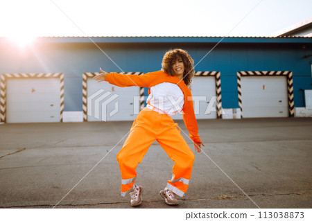 Young African American woman - dancer dancing in the street at sunset. Stylish woman with curly hair in an orange suit showing some moves Young African American woman - dancer dancing in the street at sunset. Stylish woman with curly hair in an orange suit showing some moves 113038873