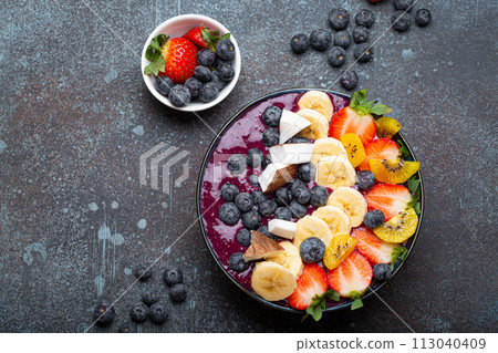 Healthy summer acai smoothie bowl with chia seeds, fresh banana, strawberry, blueberry, cocos, kiwi top view, rustic concrete background with spoon 113040409