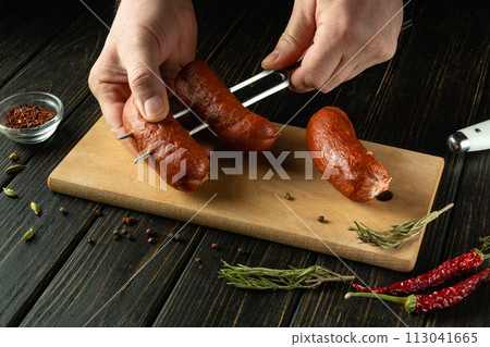 Preparing to cook a delicious grill with Munich sausages. A fork with sausages in the hands of a man on the kitchen table. Place for recipe 113041665