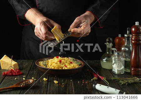 The chef grates hard cheese into a plate of pasta using a hand grater. Work environment in hotel kitchen with spices and kitchen utensils The chef grates hard cheese into a plate of pasta using a hand grater. Work environment in hotel kitchen with spices and kitchen utensils 113041666
