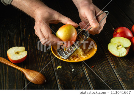 A man hands grate an apple on the kitchen table. Low key concept of preparing a healthy fruit breakfast at home A man hands grate an apple on the kitchen table. Low key concept of preparing a healthy fruit breakfast at home 113041667