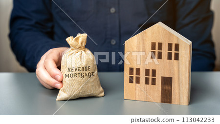 Reverse mortgage concept. Allowing homeowners to borrow money by using their home as collateral for the loan. Money bag in the hands of a businessman and a wooden house Reverse mortgage concept. Allowing homeowners to borrow money by using their home as collateral for the loan. Money bag in the hands of a businessman and a wooden house 113042233