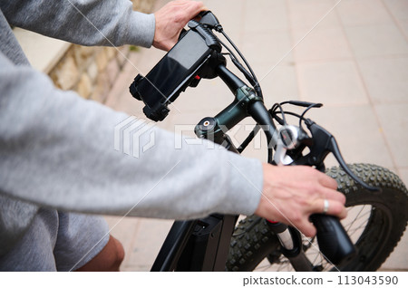 Close-up male hands on the handlebar of an electric bike in the city, smartphone with black mockup screen copy ad space Close-up male hands on the handlebar of an electric bike in the city, smartphone with black mockup screen copy ad space 113043590