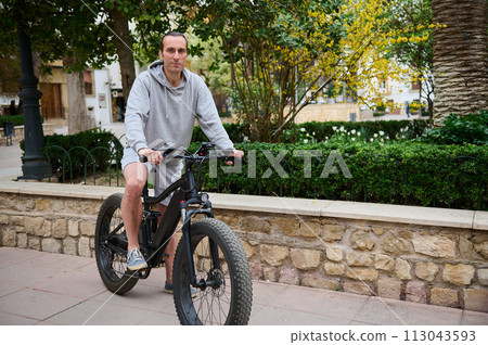 Full length portrait of a handsome Caucasian man renting electric bike, riding in the city. Bike sharing city service 113043593