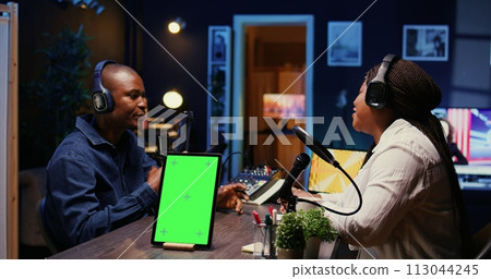 Isolated screen tablet next to vlogger show host chatting with guest in neon lights ornate living room studio. Chroma key device on table with man and woman sitting and discussing on podcast 113044245