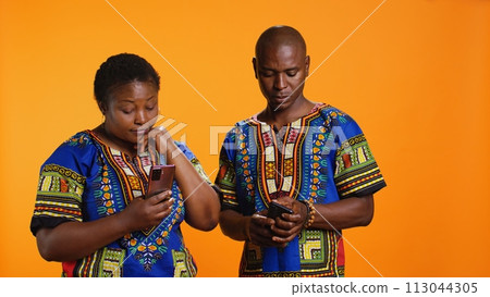 Ethnic people in traditional clothes checking smartphone apps, looking at messages received on social media network. African american man and woman browsing websites on mobile device. Ethnic people in traditional clothes checking smartphone apps, looking at messages received on social media network. African american man and woman browsing websites on mobile device. 113044305