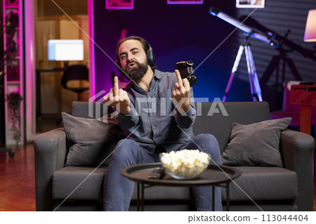 Happy gamer releasing pent up emotions after finally defeating enemies, showing middle finger sign. Man congratulating himself after winning videogame, feeling relieved, doing vulgar hand movements 113044404