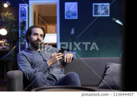 Man lounged on couch texting friend on smartphone, inviting him to watch movie on streaming service. Person at home discussing with mate on mobile phone, recommending TV show Man lounged on couch texting friend on smartphone, inviting him to watch movie on streaming service. Person at home discussing with mate on mobile phone, recommending TV show 113044407