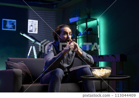 Man watching shows on TV and eating popcorn, reacting with surprise to plot twist, paying close attention to screen. Man sitting on couch in apartment shocked by film unexpected turn of events Man watching shows on TV and eating popcorn, reacting with surprise to plot twist, paying close attention to screen. Man sitting on couch in apartment shocked by film unexpected turn of events 113044409