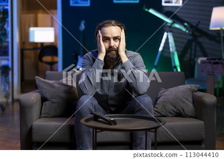 Man gasping while watching horror movie on streaming service, enjoying time off from work. Person covering mouth in disbelief, surprised by film scene broadcasted on TV set 113044410