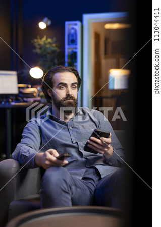 Man holding remote control while watching interesting movie on streaming service, enjoying time off from work. Person on couch surprised by high quality film broadcasted on TV 113044411