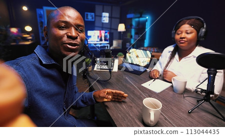 Man recording podcast using selfie camera on cellphone and high tech microphones to interview guest, ensuring flawless video and sound quality for audience watching at home, POV shot 113044435