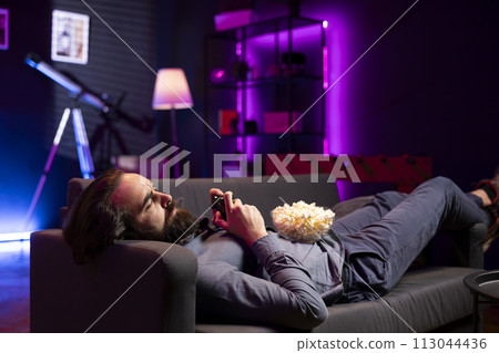 Gamer lounged on couched while engaging in online competitive multiplayer tourney using gaming console and gamepad. Man in home theatre playing videogames with bowl of popcorn in lap Gamer lounged on couched while engaging in online competitive multiplayer tourney using gaming console and gamepad. Man in home theatre playing videogames with bowl of popcorn in lap 113044436